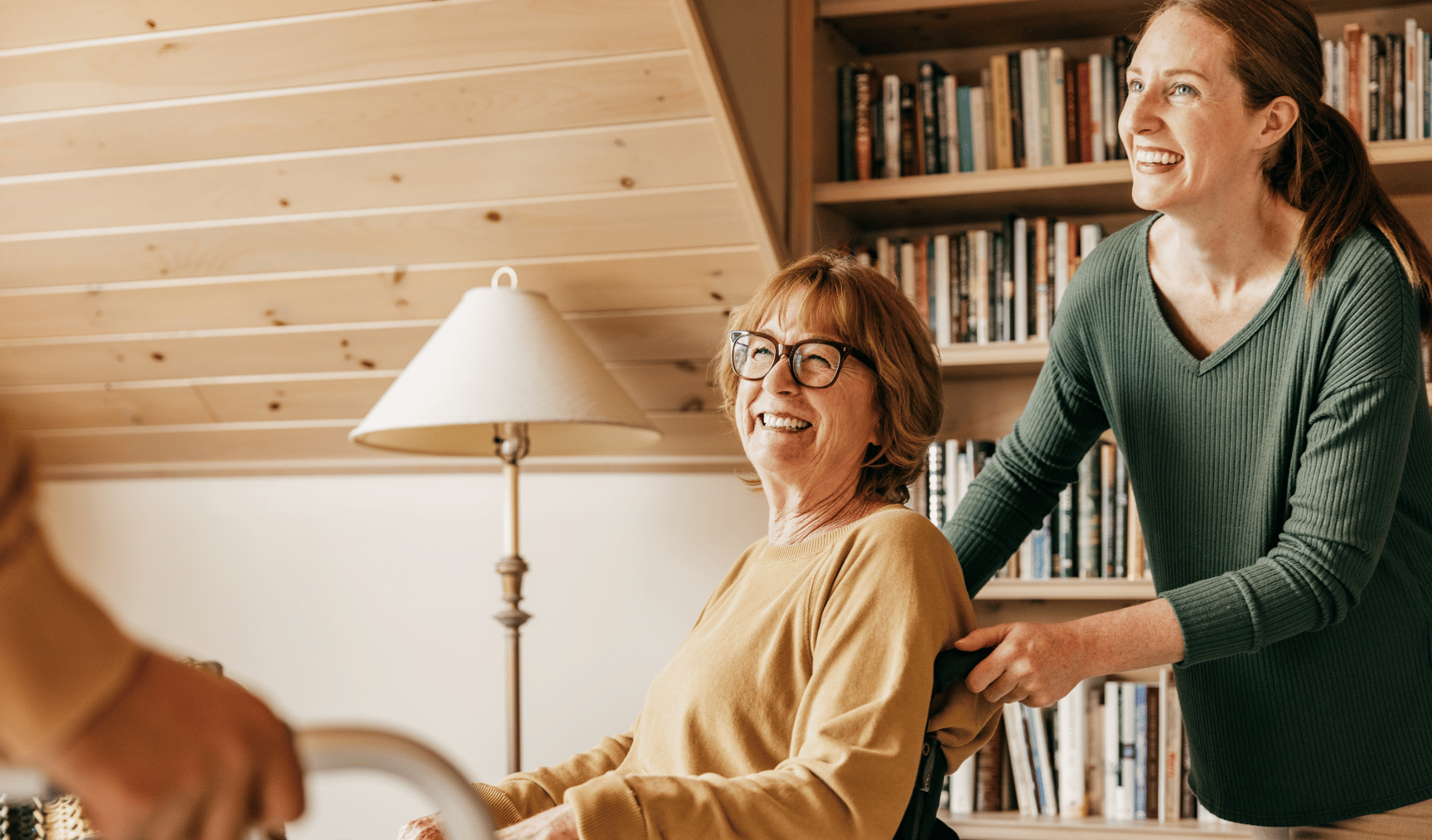 Smiling woman in glasses seated in a wheelchair, being assisted by another smiling woman in a cozy room with bookshelves in the background.