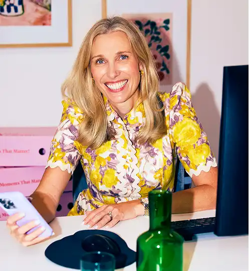A smiling woman with long blonde hair sits at a desk, holding a smartphone. She wears a floral yellow dress. The mood is cheerful and professional.