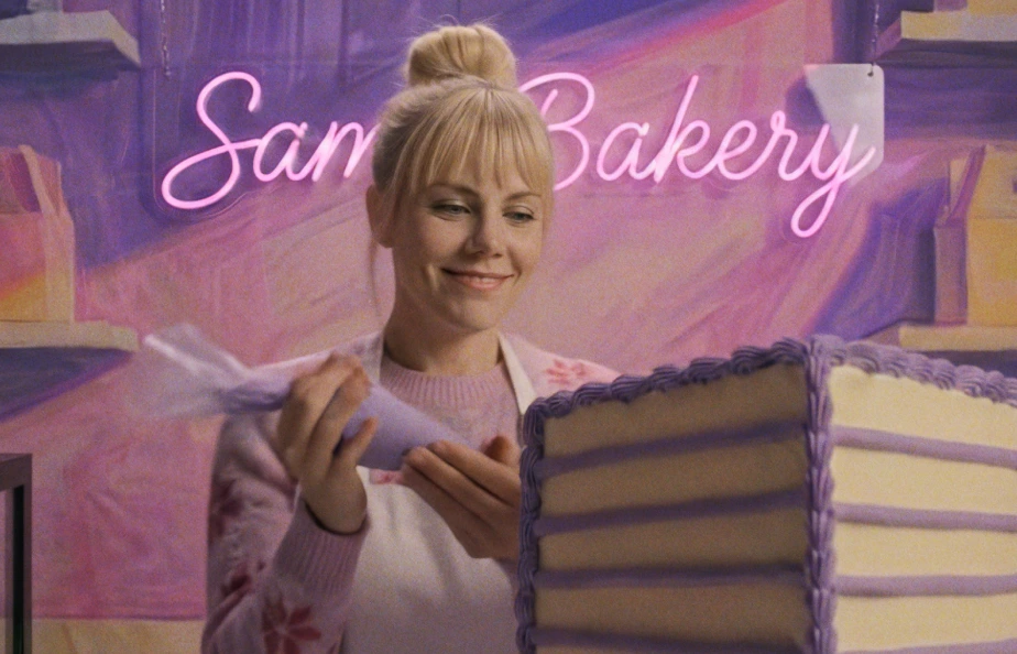 A woman with blonde hair decorates a cake with purple icing, smiling contentedly. Behind her, a neon sign reads "Sam's Bakery," set against a pastel background.