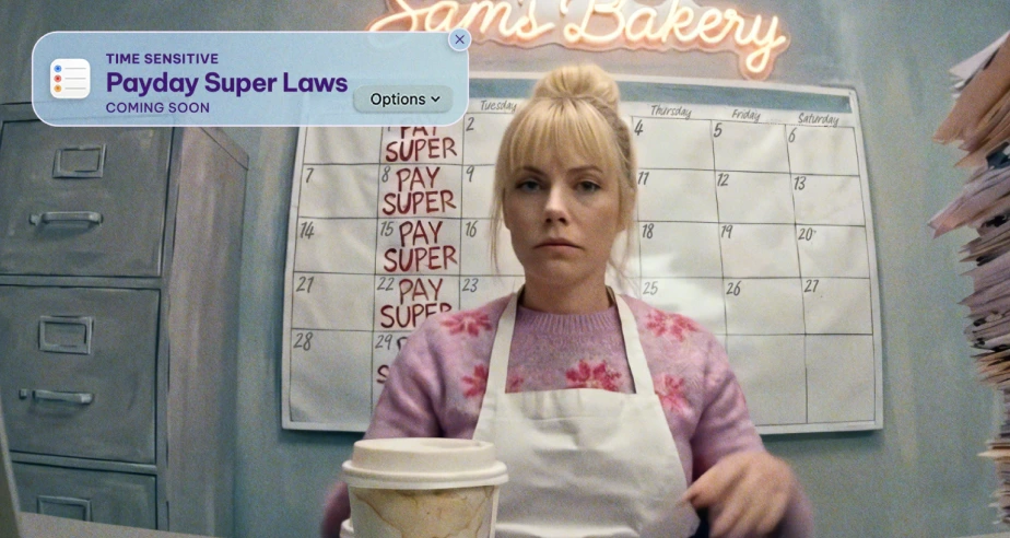 A woman sits at a desk in front of a wall calendar. She is wearing a pink sweater and white apron, looking serious. A coffee cup is on the table. A phone notification about "Payday Super Laws" is visible. The setting appears to be a bakery, as a neon "Sam's Bakery" sign is partially visible above. Stacks of papers are on the right, suggesting busyness or paperwork.