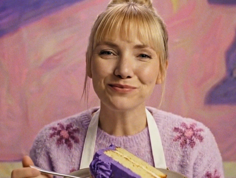 A woman in a purple sweater, with a white apron, smiles warmly, holding a slice of cake with purple frosting. The background is soft, painted pastel colors.