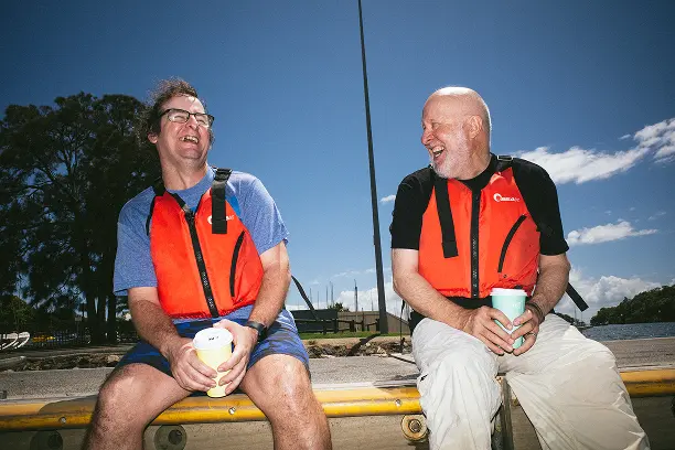 Two men sit on a dock, wearing red life vests, laughing and holding cups. The sunny sky and trees create a joyful, relaxed atmosphere.