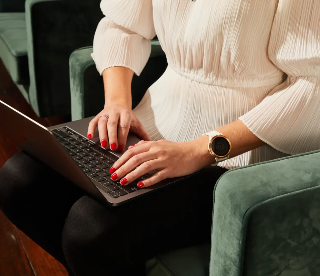 A woman with red nail polish types on a laptop while seated on a green velvet chair, wearing a white blouse and smartwatch.